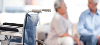 Florida Assisted Living Association. Two people in the background sitting, out of focus. In the foreground on the left is an empty wheelchair.