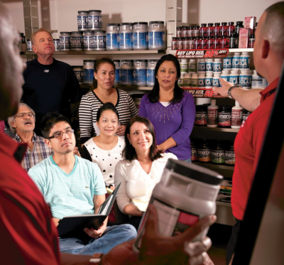Two Max Muscle employees talk to a group of people with a board behind them and product in hand.