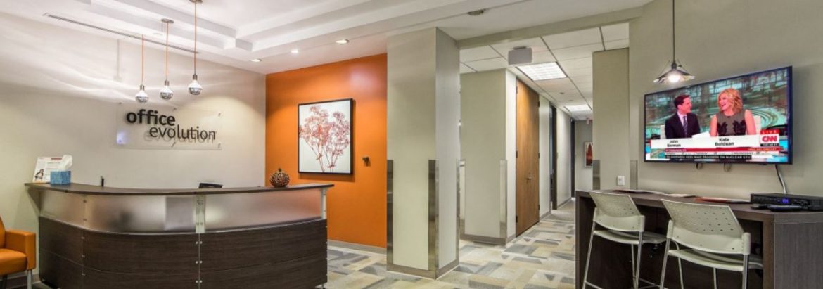 Office Evolution office with their sign on the left by a reception counter and a tv to the right of a hall on the right.