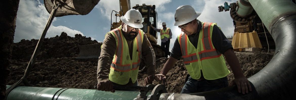 Two menu in hard hats and reflective vests look over a pipe connection with a worker behind them at a crane.