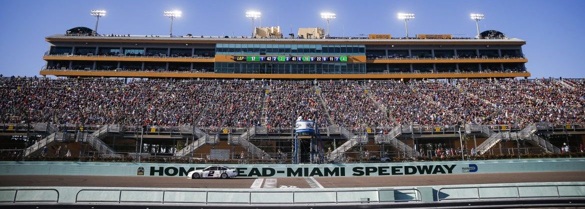 Homestead-Miami Speedway, a view of the main speedway building full of fans with lights on and a dimming blue sky behind.