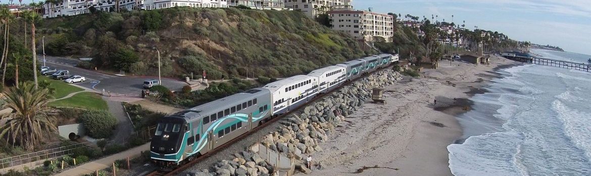 Metrolink, a train travelling on tracks along a beach with a hill and multi story buildings behind.