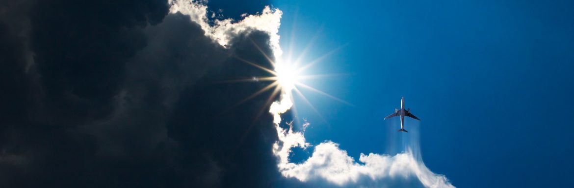 Roswell International Air Center. A jet flying through clouds making a trail with the sun peaking out of dark clouds on the left.