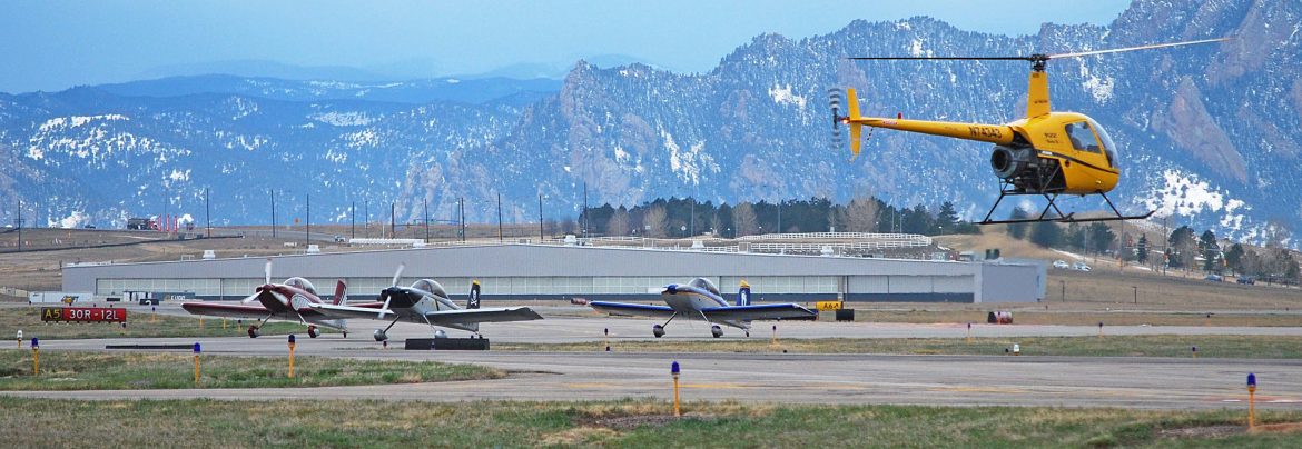 Rocky Mountain Metropolitan Airport a helicopter in flight with airplanes on the ground beyond, mountains in the background.