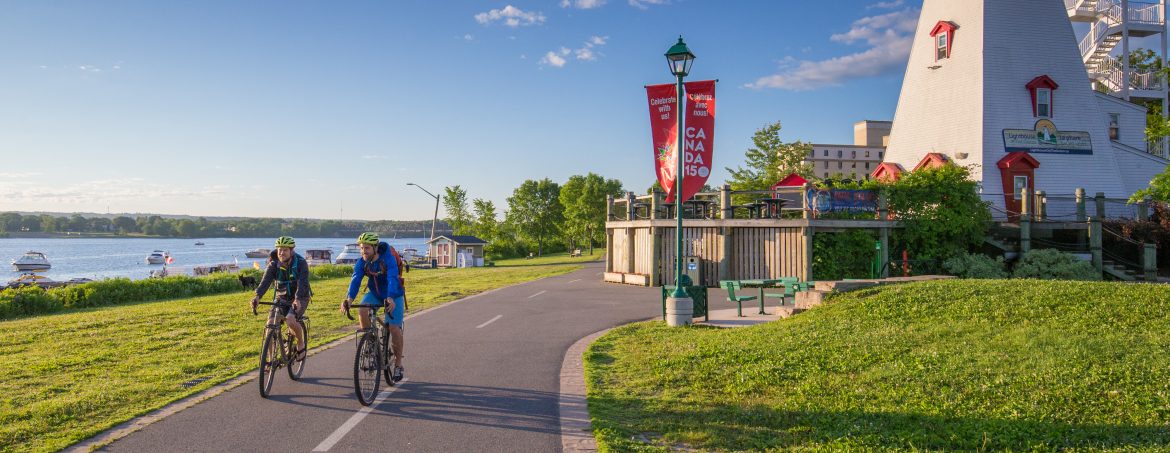 Fredericton, New Brunswick. Cyclers on a bike path along the water and a lighthouse on the right.