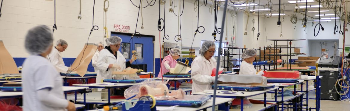 AIM Aerospace production lines with multiple people wearing hair nets at workstations with cables hanging down from the ceiling powering tools.
