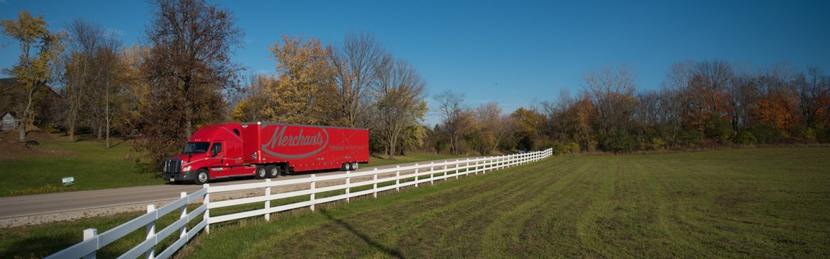 Merchants Moving and Storage semi truck traveling through a green field on a road with a white fence and trees behind.