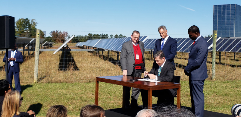 The North Carolina Building Performance Association event with a man sitting at a table in a grassy area with a solar array behind him. Gentlement in suits standing behind him and a few heads visible in the foreground from people sitting and watching.