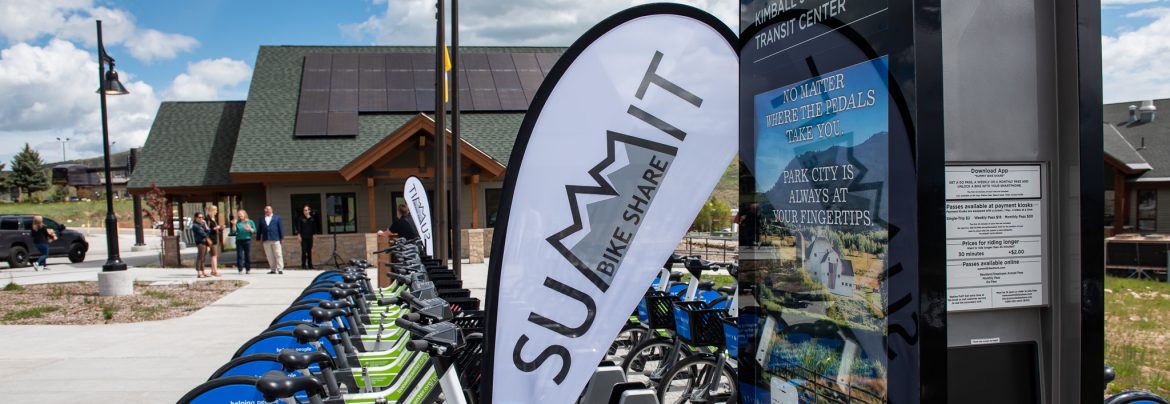 Summit County, Utah Summit bike share sign with bikes lined up in a row behind and a building with solar panels on the roof in the background.
