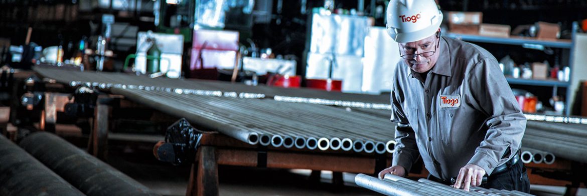 Tioga Pipe, Inc. a man with a helmet on inspecting pipes on a table with pipes behind in a warehouse.