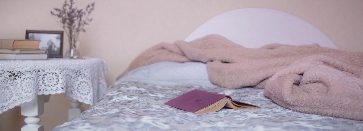 A bed and nightstand in a room with a blanket bunched up on the bed and a book.