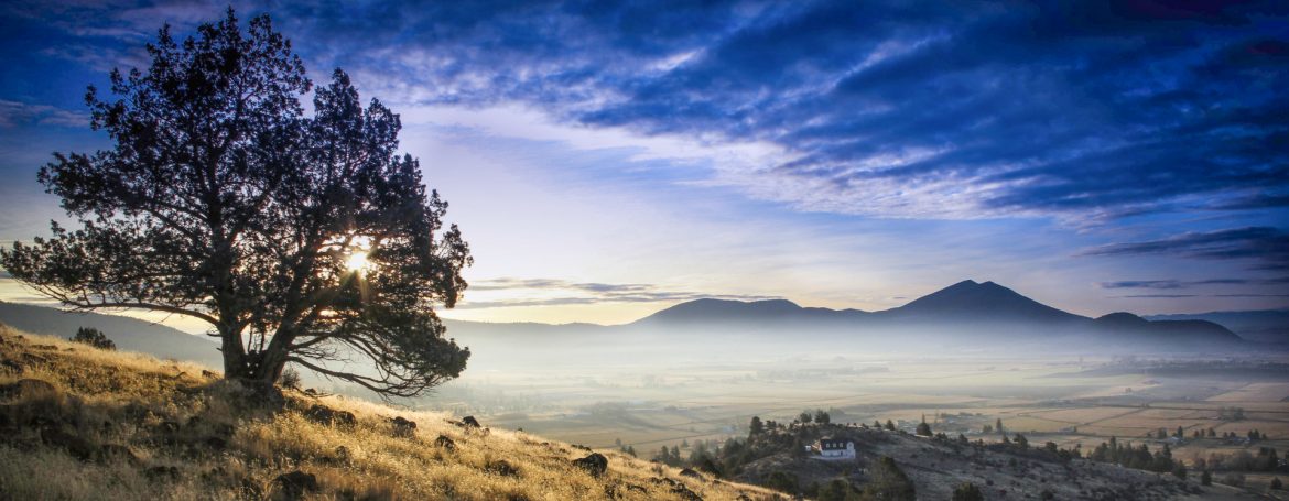 Klamath County, Oregon. Beautiful photo of blue sky and clouds while look at a hill on the left with a tree and to the right beyond is a lower lying area with a house and fog.