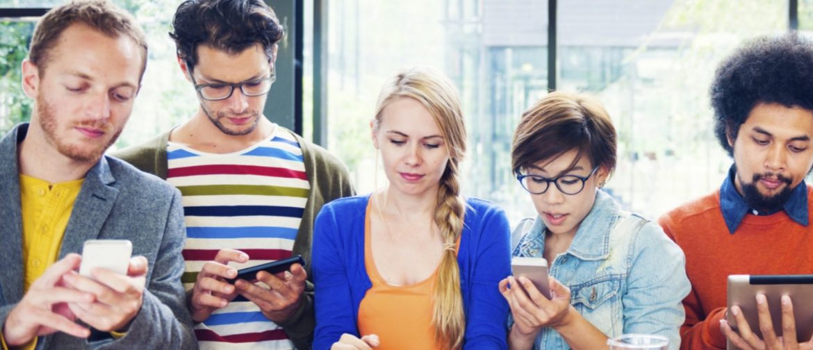 5 young men and women are in a row at a table all holding tablets or phones, looking down and using them.