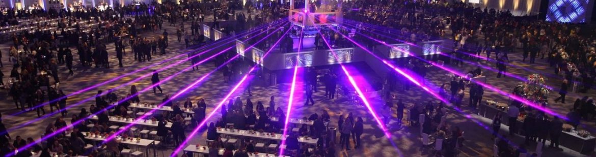 The Professional Convention Management Association; A view of a convention with lights shining out over the room in beams.