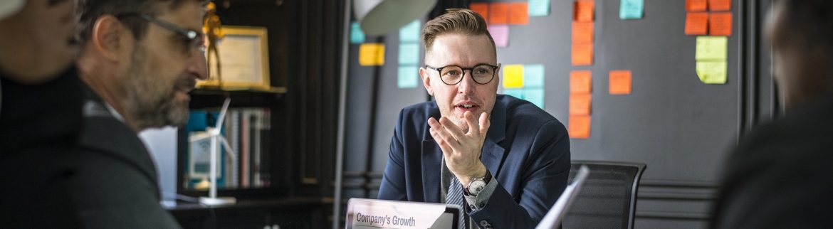 Business people talking at a desk with a computer in front of one person and sticky notes on the wall behind.