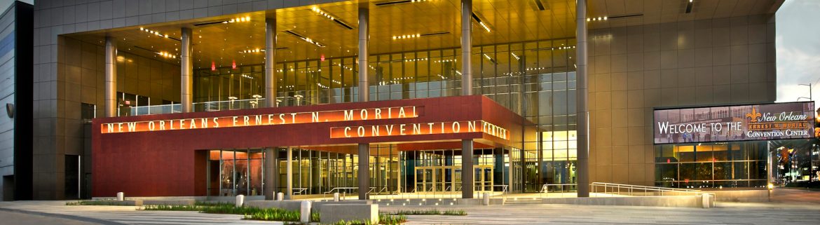 New Orleans Ernest N. Morial Convention Center front entrance with some daylight left but lights on and glowing. Cloud and blue sky visible in background.