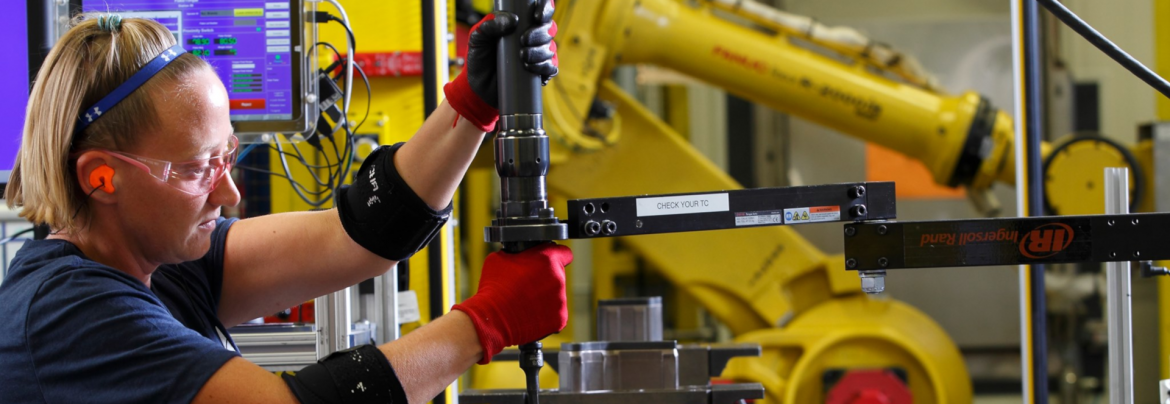 Sekisui Aerospace, formerly AIM Aerospace. A woman using a tool on a part in a factory / warehouse setting with robotic arms in the background..