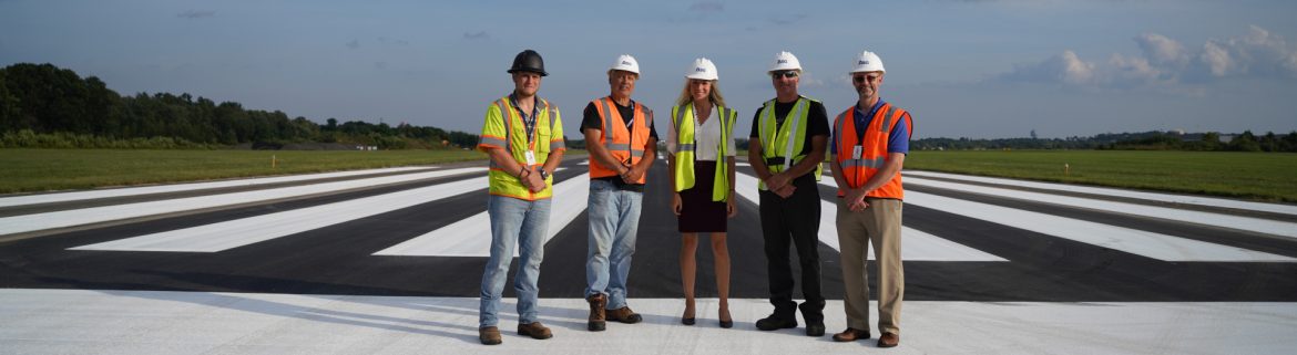 Beverly Regional Airport group photo on the runway. 5 people with hard hats and reflective vests on.