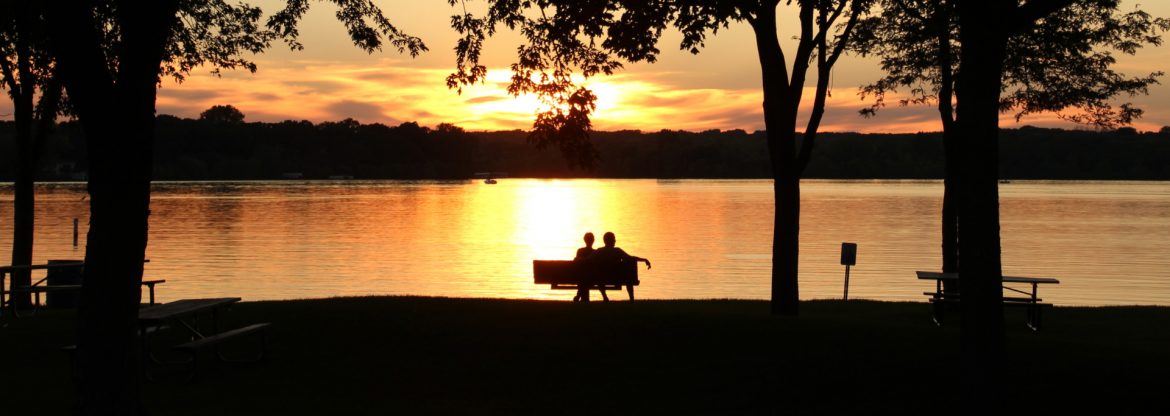 Lakeville, Minnesota Antlers park sunset with dark black foreground shapes and a bench with two adults sitting in front of the suns reflection on the water.