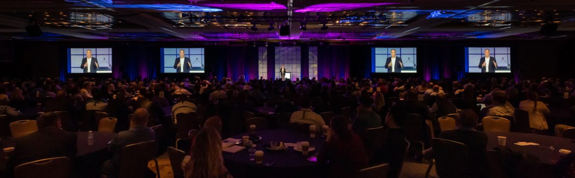 The Association for Manufacturing Excellence - AME event room with people sitting at tables, monitors in the background and speakers on stage.